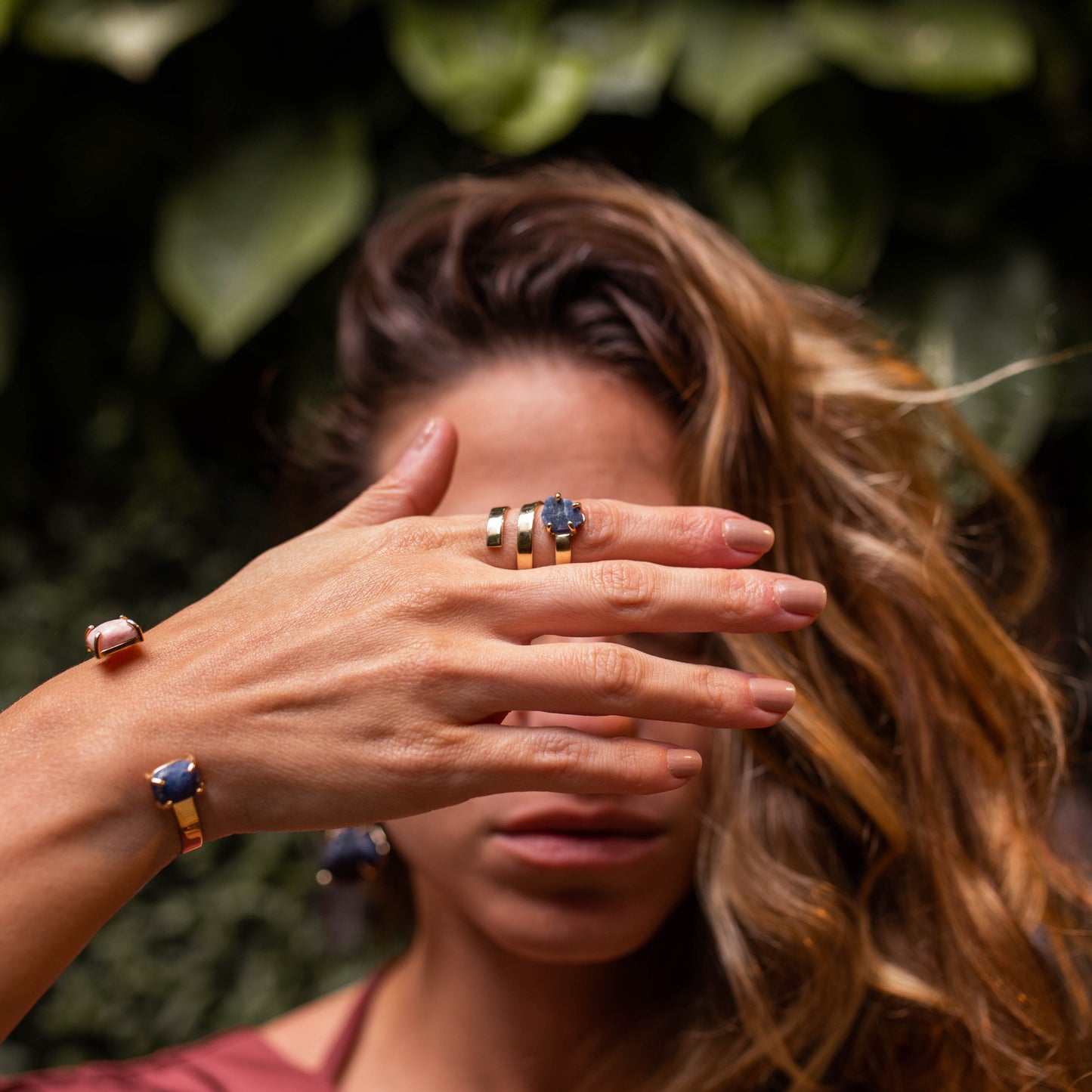 Mulher tapando o rosto com a mão usando anel e pulseira Ushuaia em ouro 18k com pedra Rodocrosita e Sodalita.
